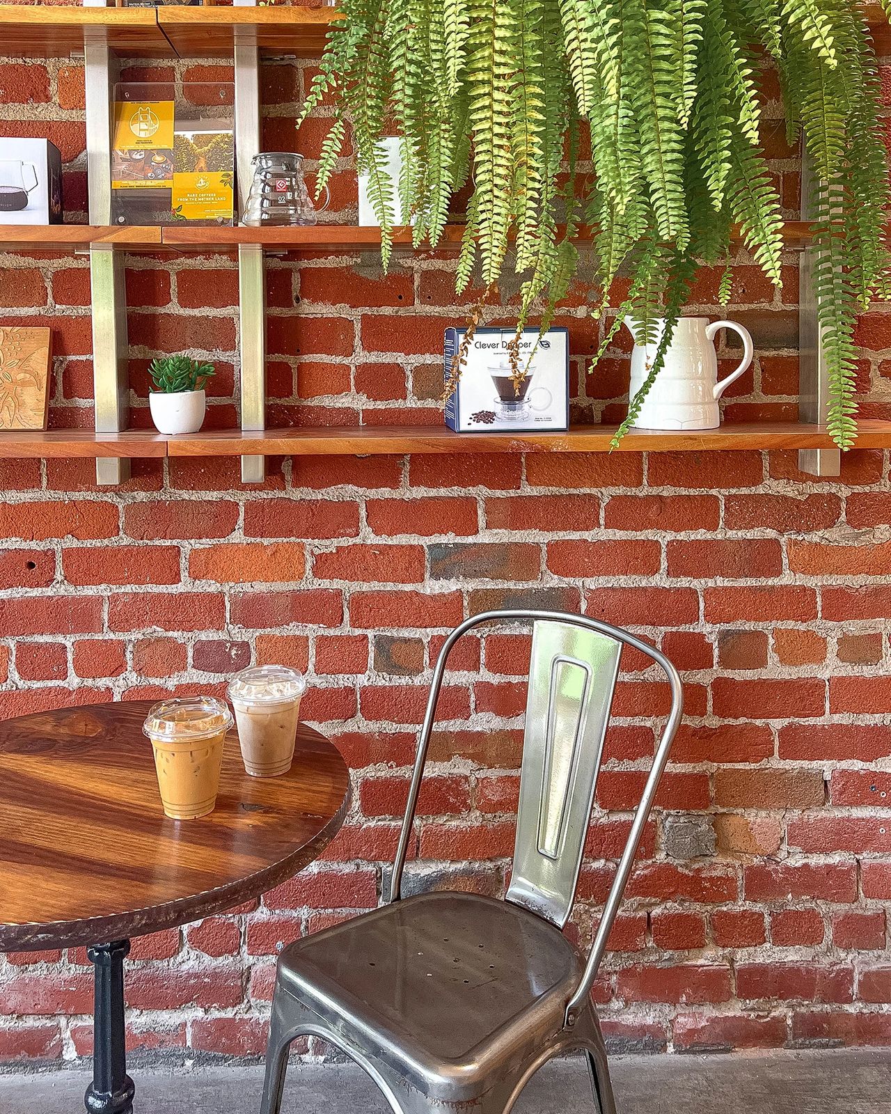 Two iced coffees on a wooden table with a metal chair and brick wall at Sextant Coffee Roasters in San Francisco, decorated with greenery and wooden shelves