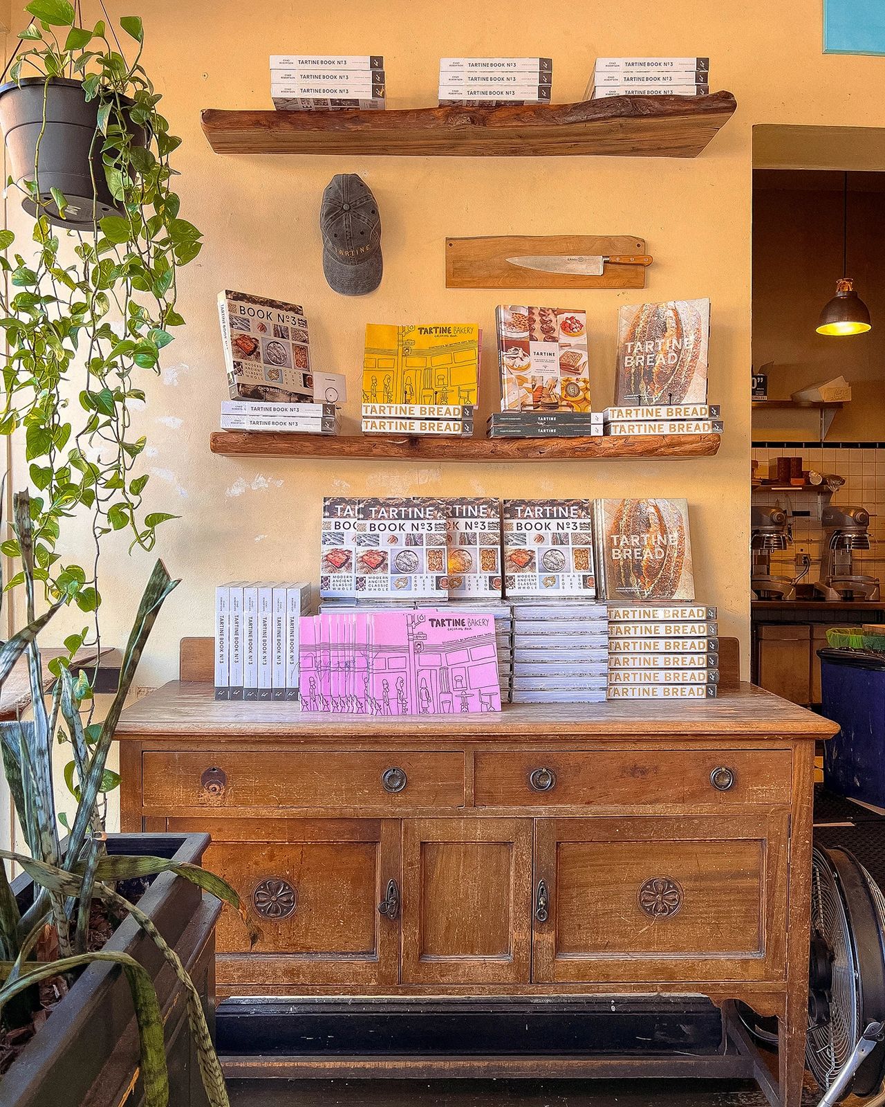 nside Tartine Bakery in San Francisco, featuring a cozy corner display of cookbooks, rustic wooden furniture, leafy plants, and a glimpse into the bakery's warm, artisanal atmosphere. 