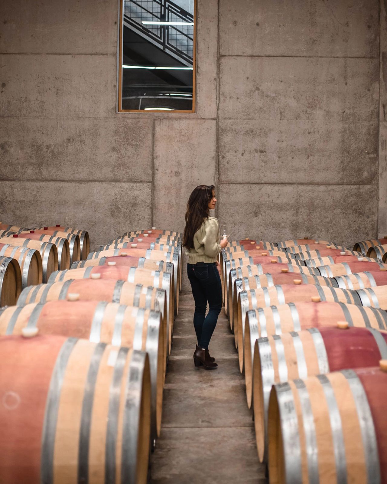 A woman walks through rows of oak wine barrels inside a modern winery cellar in Oregon’s Willamette Valley. The concrete walls, stacked barrels, and warm lighting create a rustic yet sophisticated atmosphere. The Willamette Valley is one of the premier wine regions in the United States, known for its world-class Pinot Noir and scenic tasting experiences just outside of Portland. Photo by Nastasia Yakoub