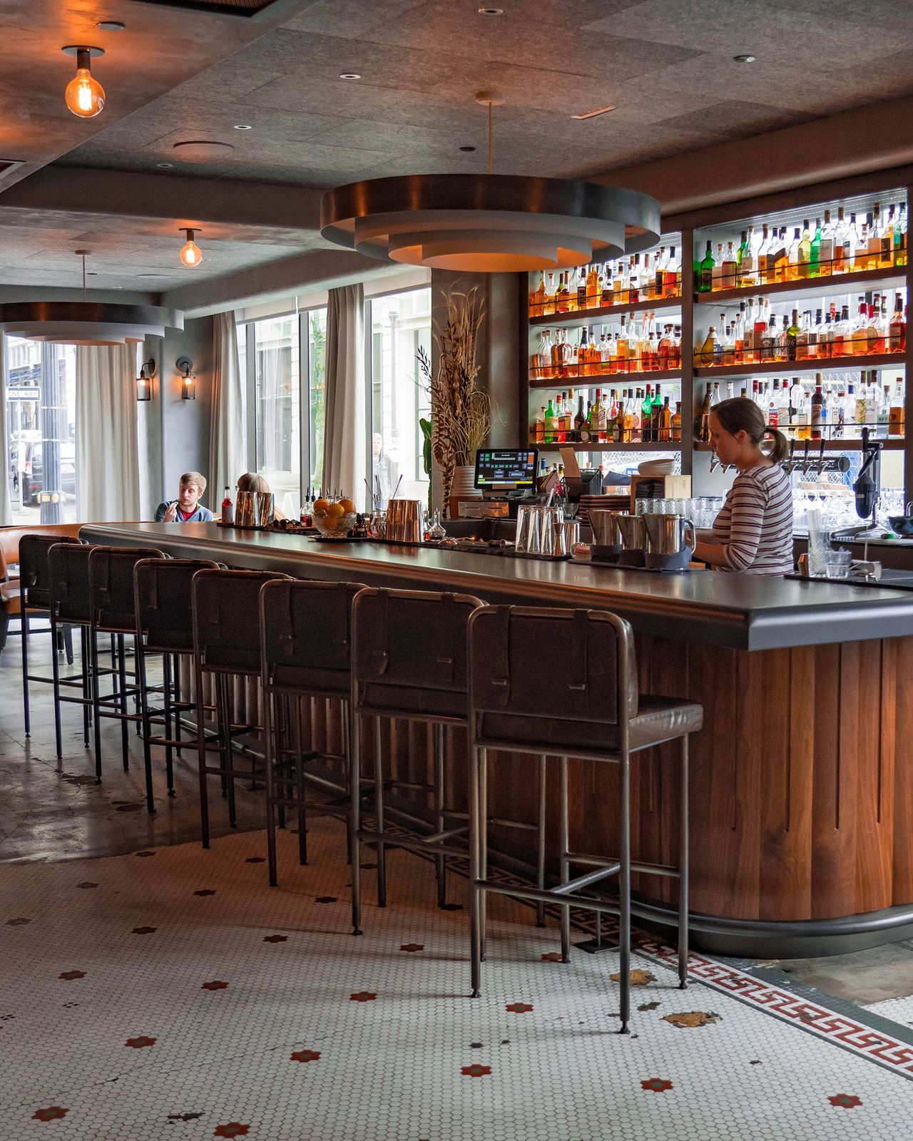 The elegant bar inside Bullard Tavern in Portland, Oregon, featuring a curved wooden bar lined with leather stools, backlit shelves stocked with colorful bottles, and soft, modern lighting. The space blends vintage charm with contemporary design, creating a warm and sophisticated atmosphere perfect for cocktails or a Texas-meets-Pacific-Northwest dining experience. Photo by Nastasia Yakoub