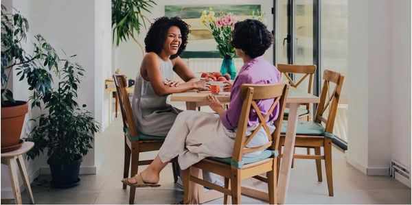 Two women happily chatting at a kitchen table with plants and flowers nearby.