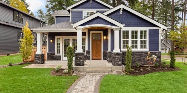 Blue house with white trim and stone accents, surrounded by green lawn and trees.