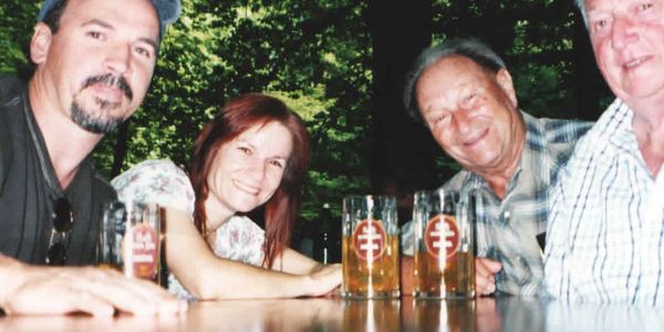 Four people smiling with glasses of beer at an outdoor table.