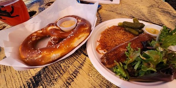 Soft pretzel with mustard and a plate with sausage, pickles, hash browns, and salad on a rustic wooden table.