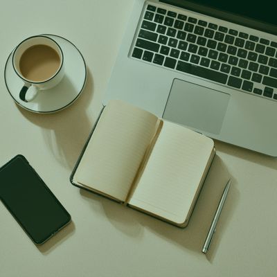 Workspace with coffee, phone, notebook, pen, and laptop on a light surface.