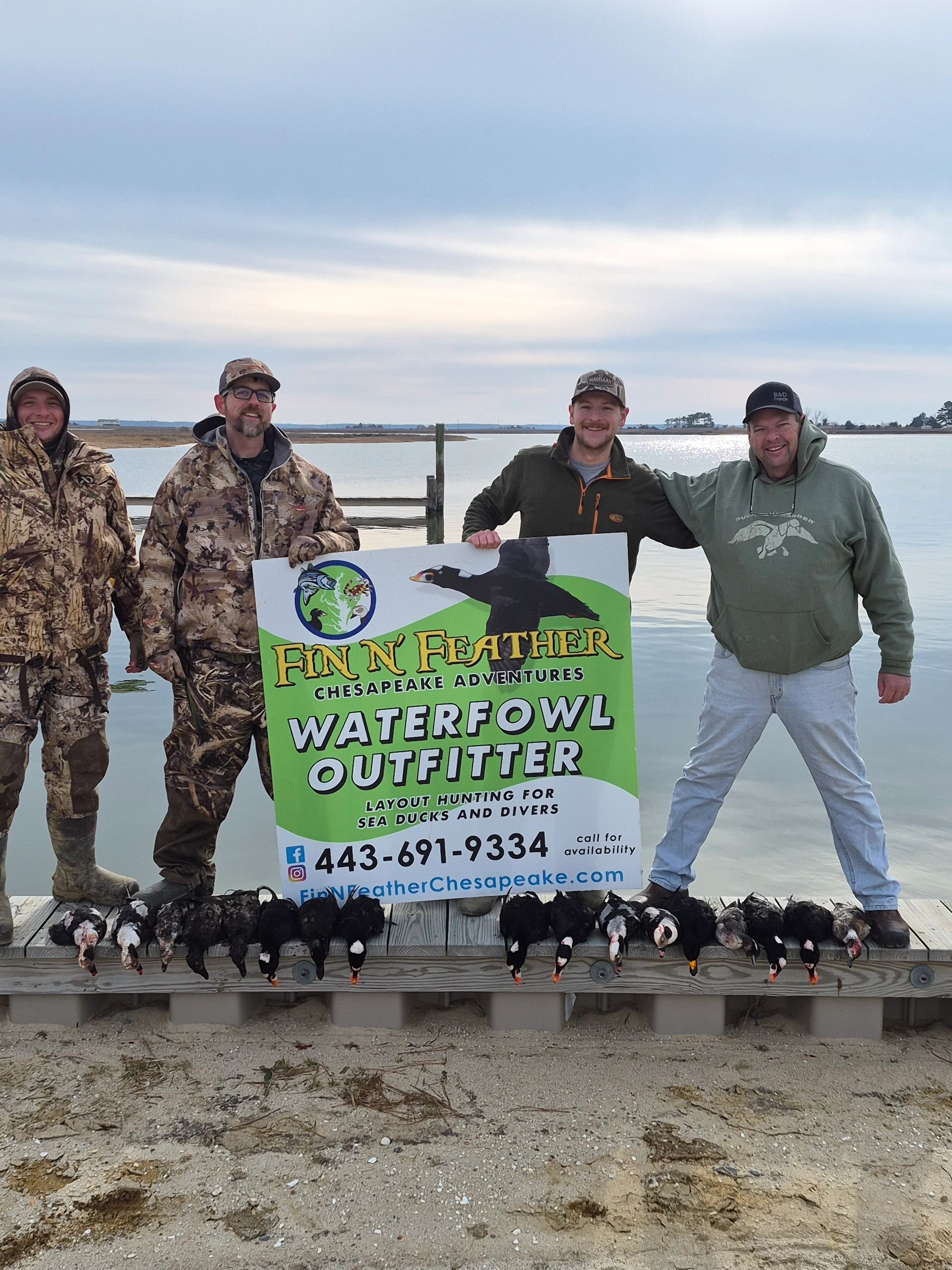 Four hunters with waterfowl and a sign for a hunting outfitter.