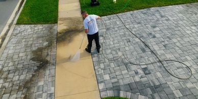 A man pressure washes a dirty sidewalk next to a driveway.