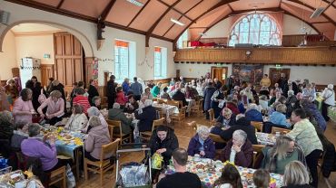 Community gathering in a large hall with tables of people eating and socializing.