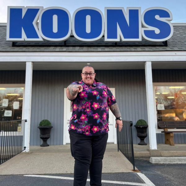 A smiling person in a floral shirt stands under a large KOONS sign.