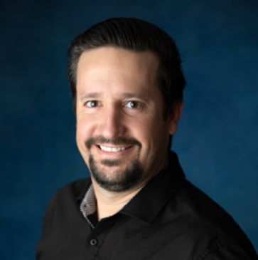 Smiling man with dark hair and beard against a blue background.