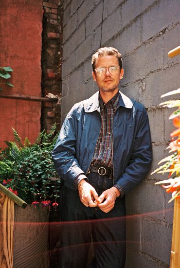 Man standing against a brick wall with greenery around.