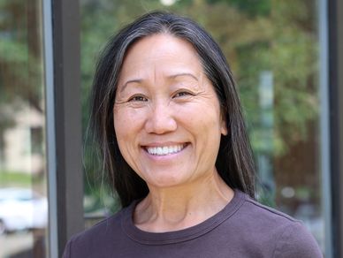 Smiling woman with long black hair standing outdoors near a window.