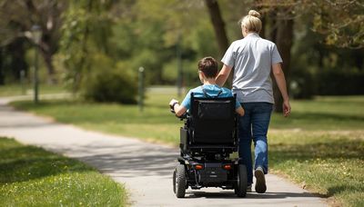 A caregiver walks with a person in a motorized wheelchair on a park path.
