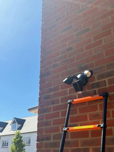 A ladder leaning against a brick wall with outdoor security lights above it.