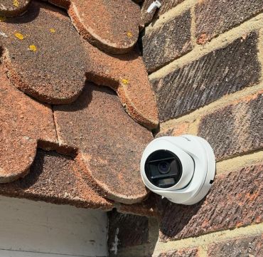 A white security camera mounted on a brick wall beneath a tiled roof.