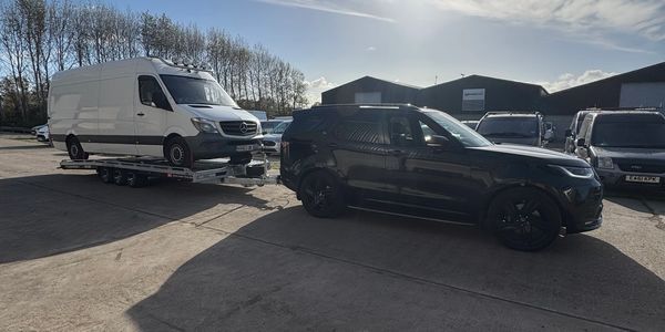 Black SUV towing a white van on a trailer in a parking lot under a bright sky.