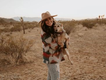 A woman in a hat holds a dog in a desert landscape.