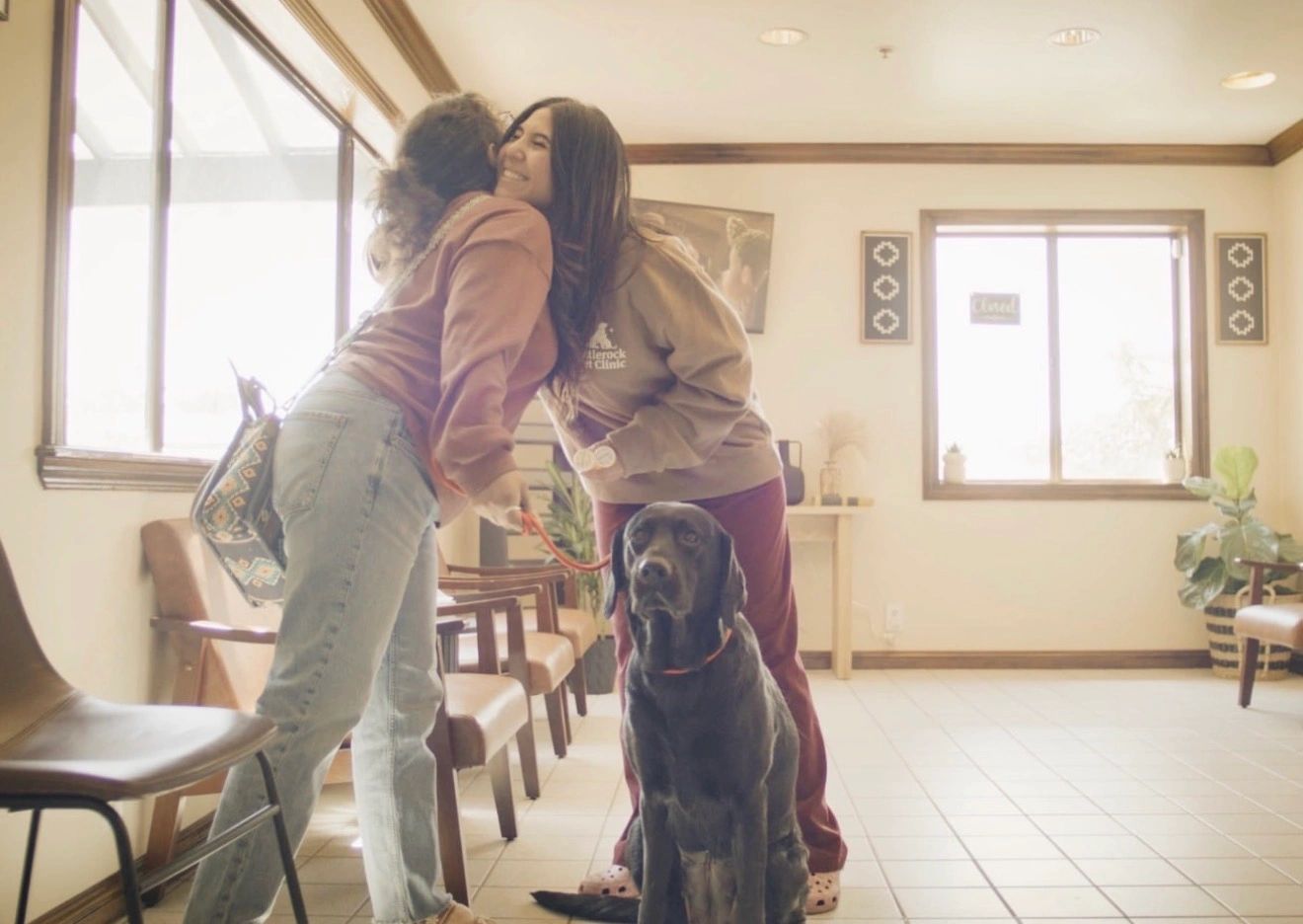 Two women warmly greeting each other with a black dog sitting nearby.