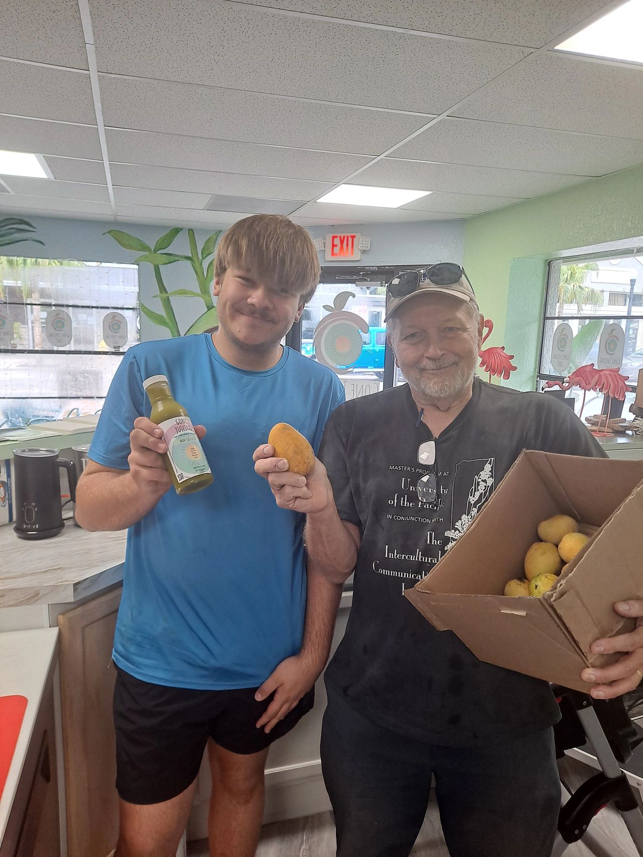 FarmNet Volunteer with delivery of fresh local mangos to Gone Juicing (New Port Richey) and staff member of Gone Juicing holding mango juice made at Gone Juicing.  Gone Juicing is a partner of NPR FarmNet.