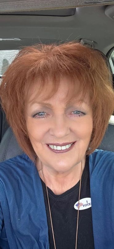 Smiling woman with red hair wearing an "I Voted" sticker in a car.