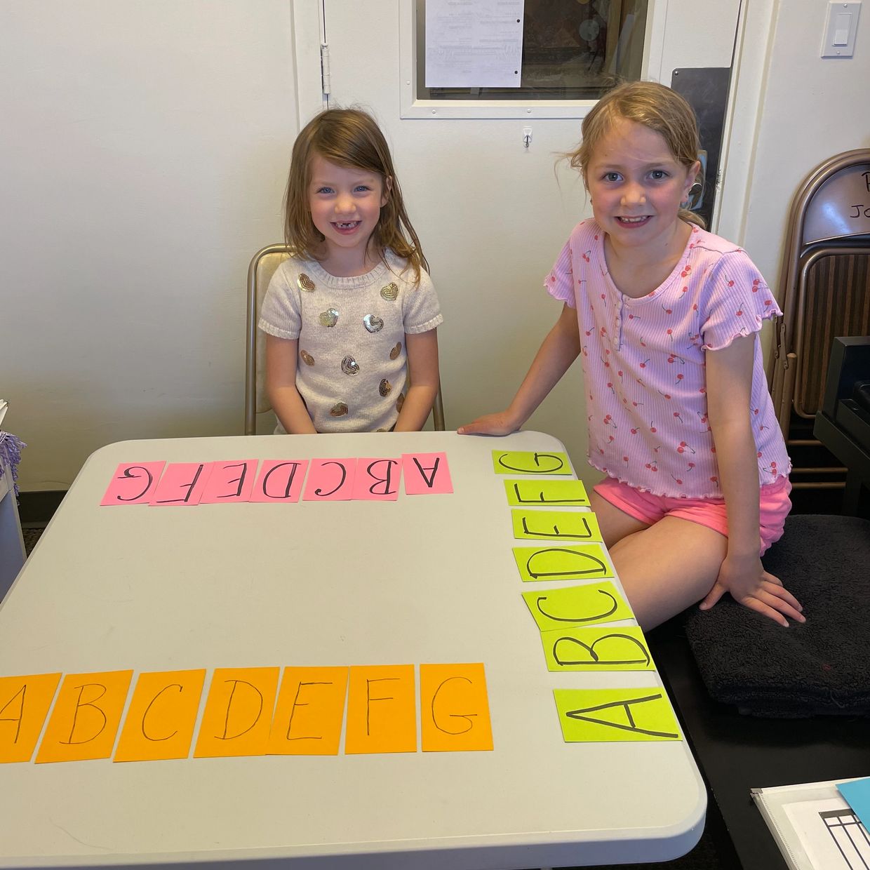 Two girls sit at a table with colorful alphabet cards arranged in rows.