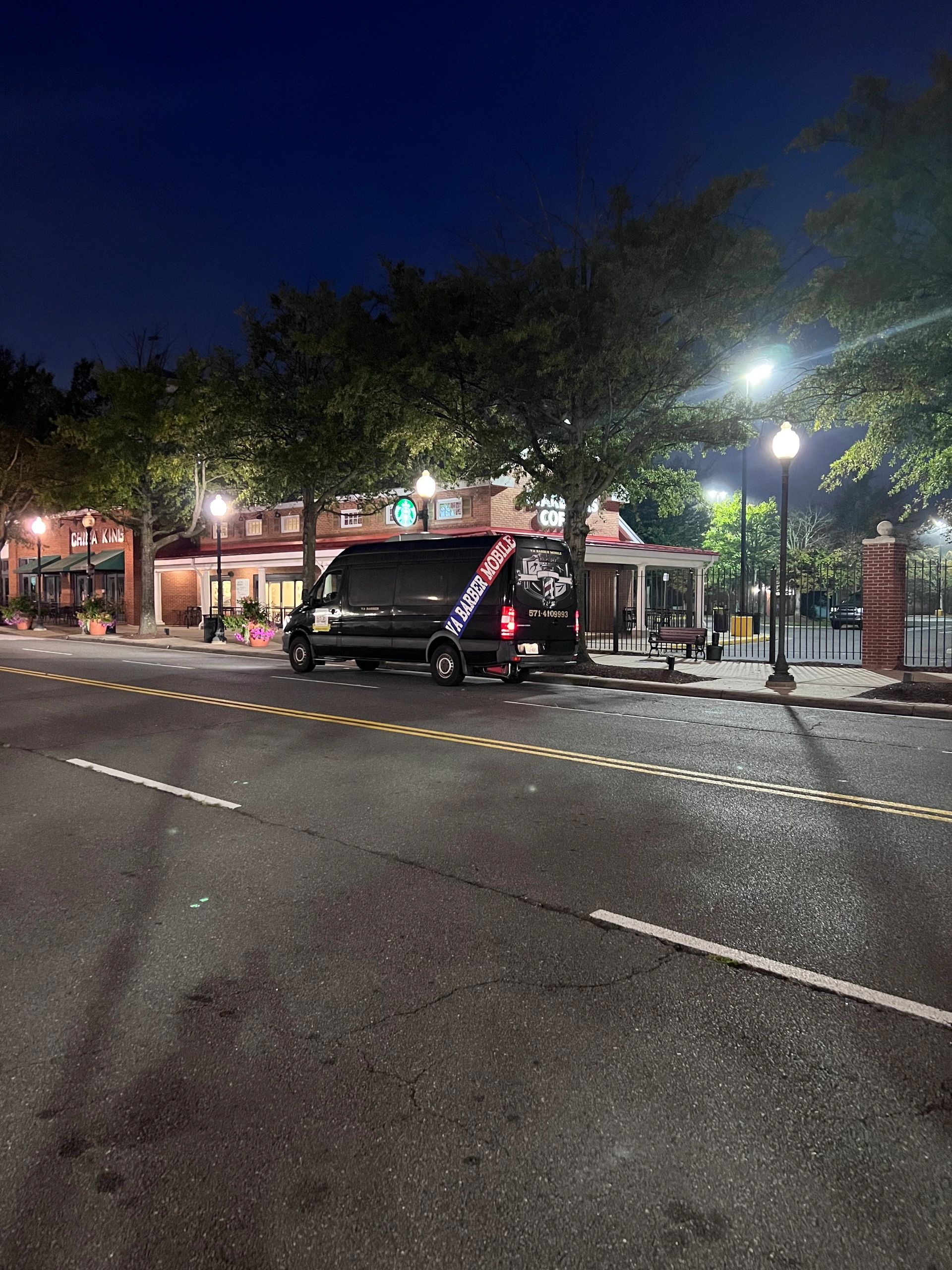 A mobile barber van parked on a quiet street at night near a Starbucks.