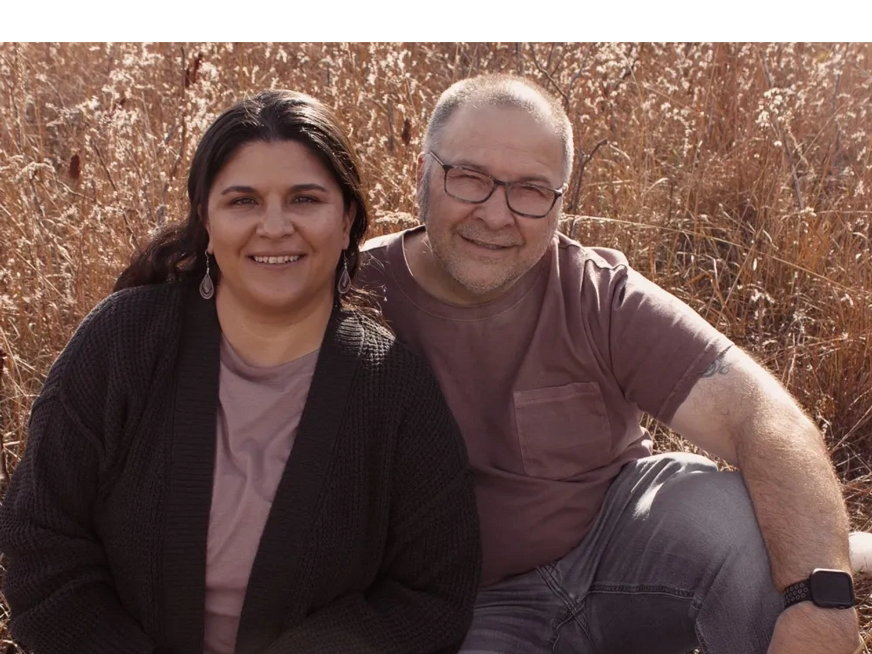 Angela and Matt sit together in a field of tall grass. 