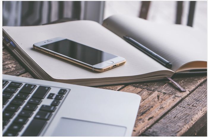 A smartphone and a pen rest on an open notebook beside a laptop