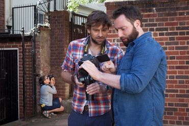 Two men reviewing photos on a camera while a woman takes pictures in the background.