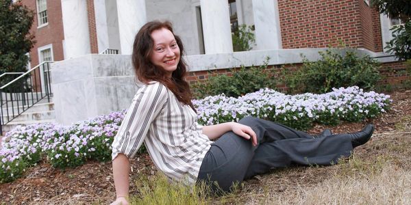 Young woman smiling while sitting on grass near flowers and a building.
