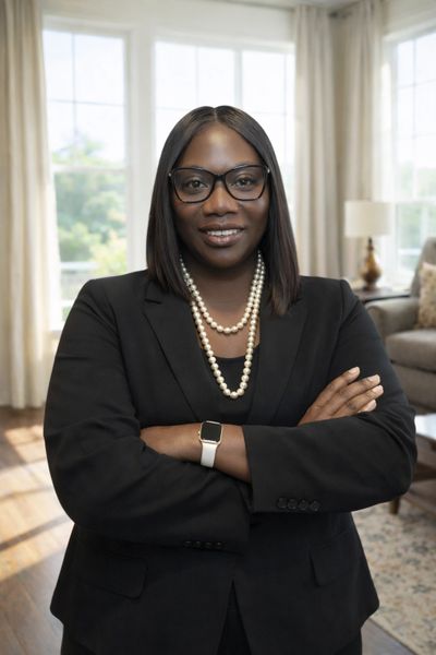 Confident woman in black blazer and pearls standing with arms crossed indoors.