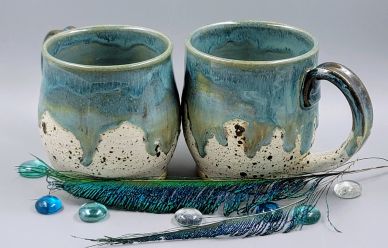 Two ceramic mugs with blue-green glaze and peacock feathers with shiny beads.
