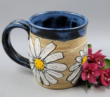 Ceramic mug with hand-painted daisies and blue rim beside pink flowers.
