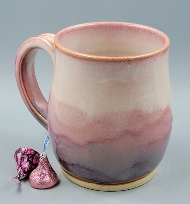 A ceramic mug with a gradient of pink, purple, and beige tones next to two wrapped chocolates.