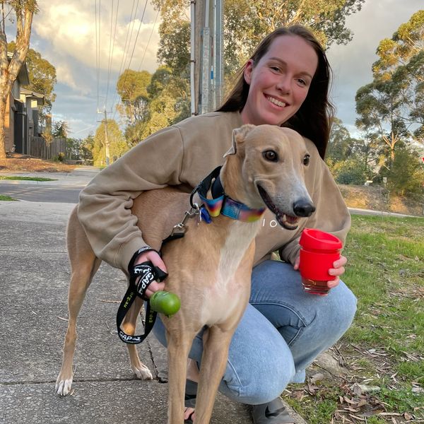 Woman with brown hair in kahki jumper and blue jeans is holding a cup of coffee next to a greyhound.