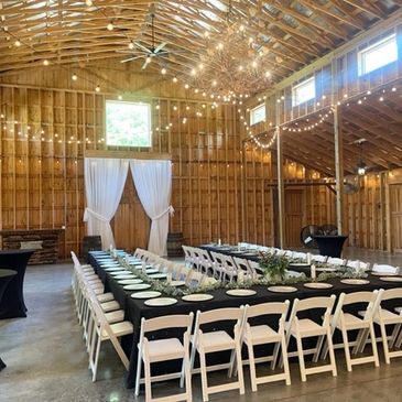Rustic barn decorated with string lights and a U-shaped dining setup for an event.