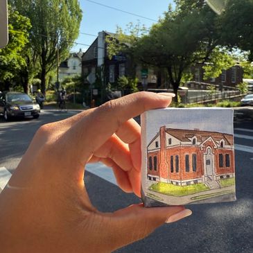 Hand holding a miniature painting of a red brick building outdoors.