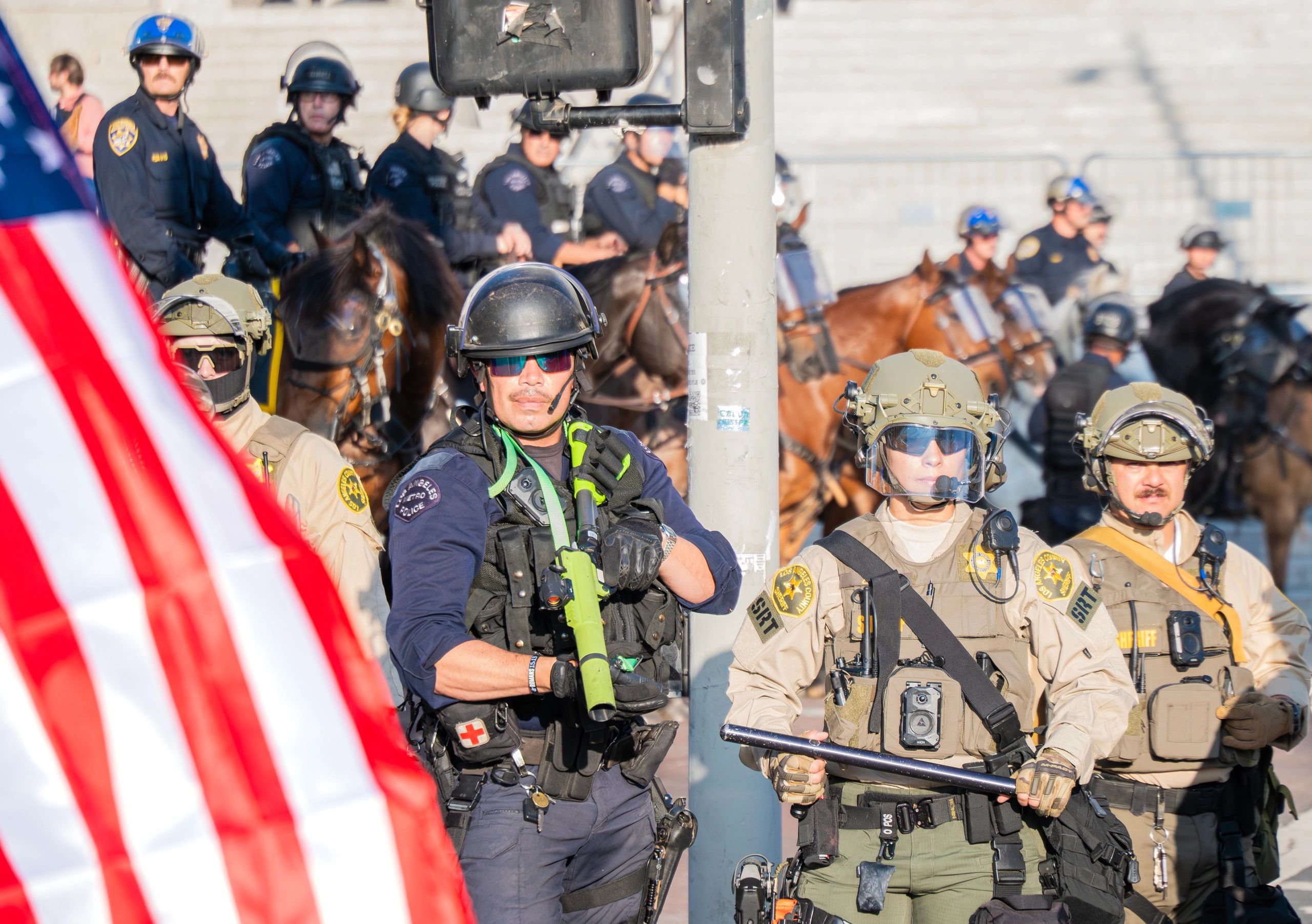 Armed law enforcement officers in tactical gear stand alert near an American flag.