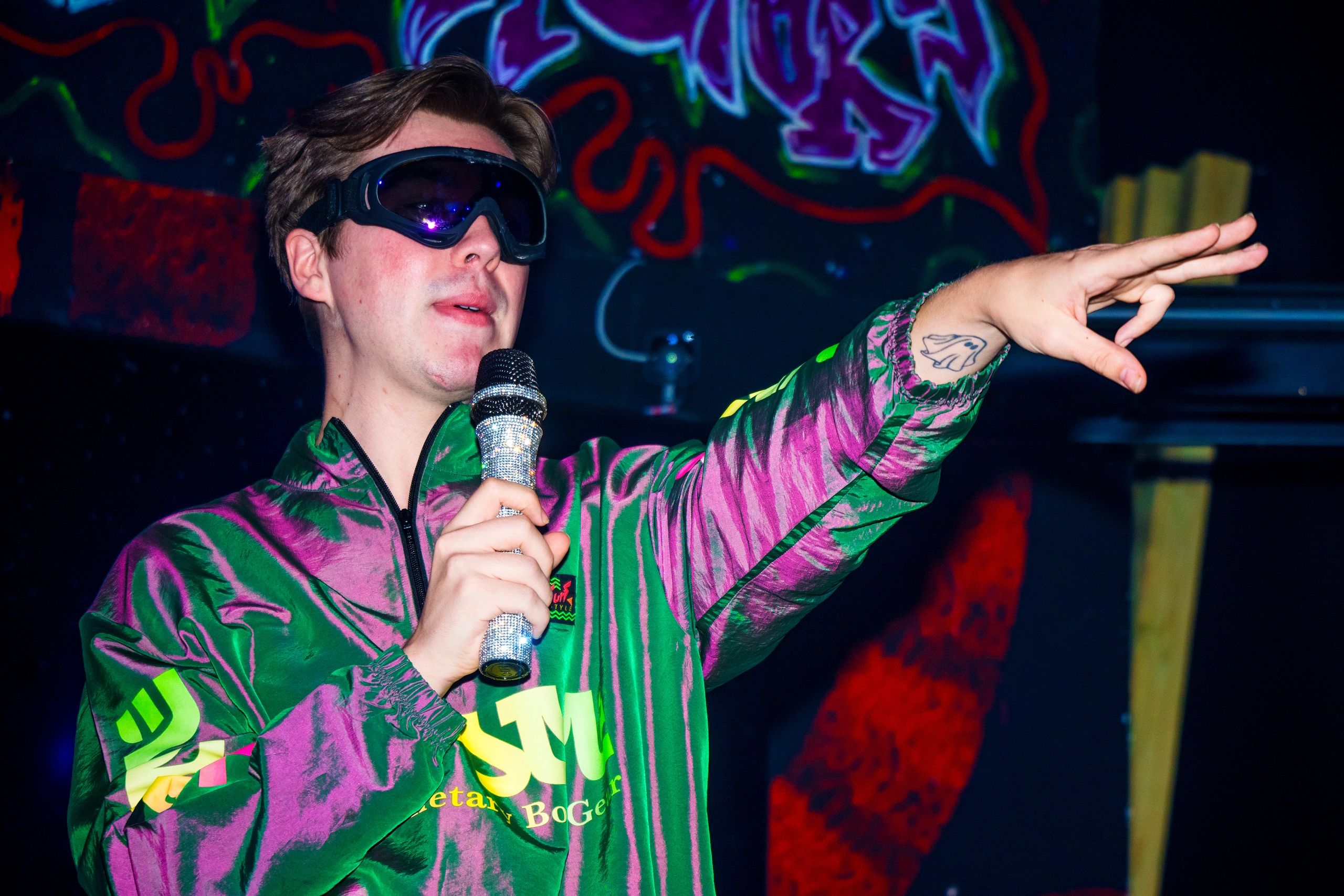 A person wearing goggles and a colorful jacket holding a microphone and pointing.