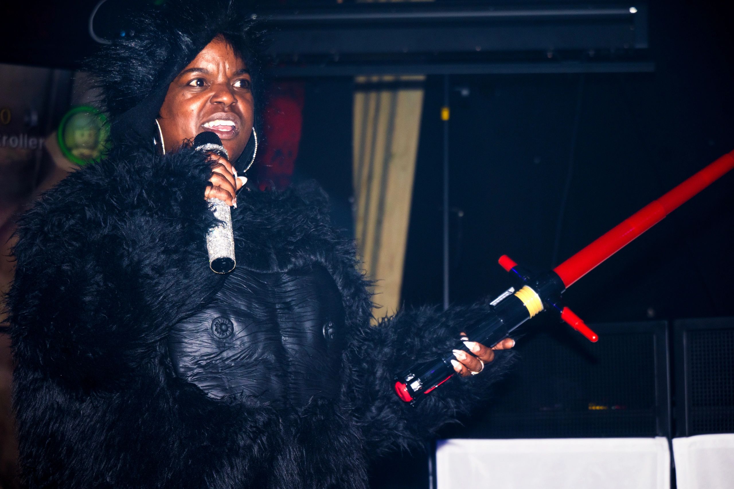 A woman in a black furry outfit holds a microphone and a red toy lightsaber.