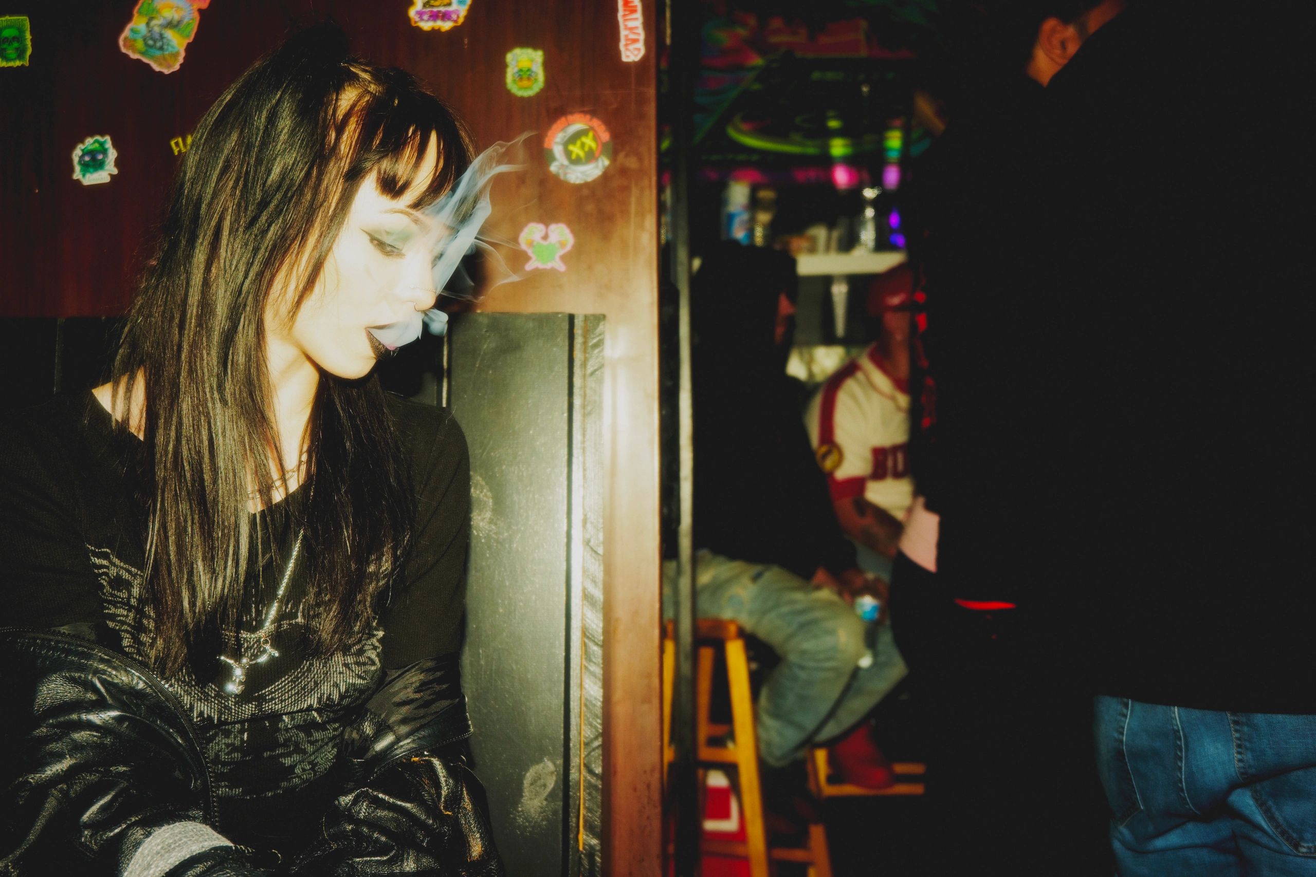 Woman in dark attire exhales smoke in a dimly lit room with stickers on the wall.