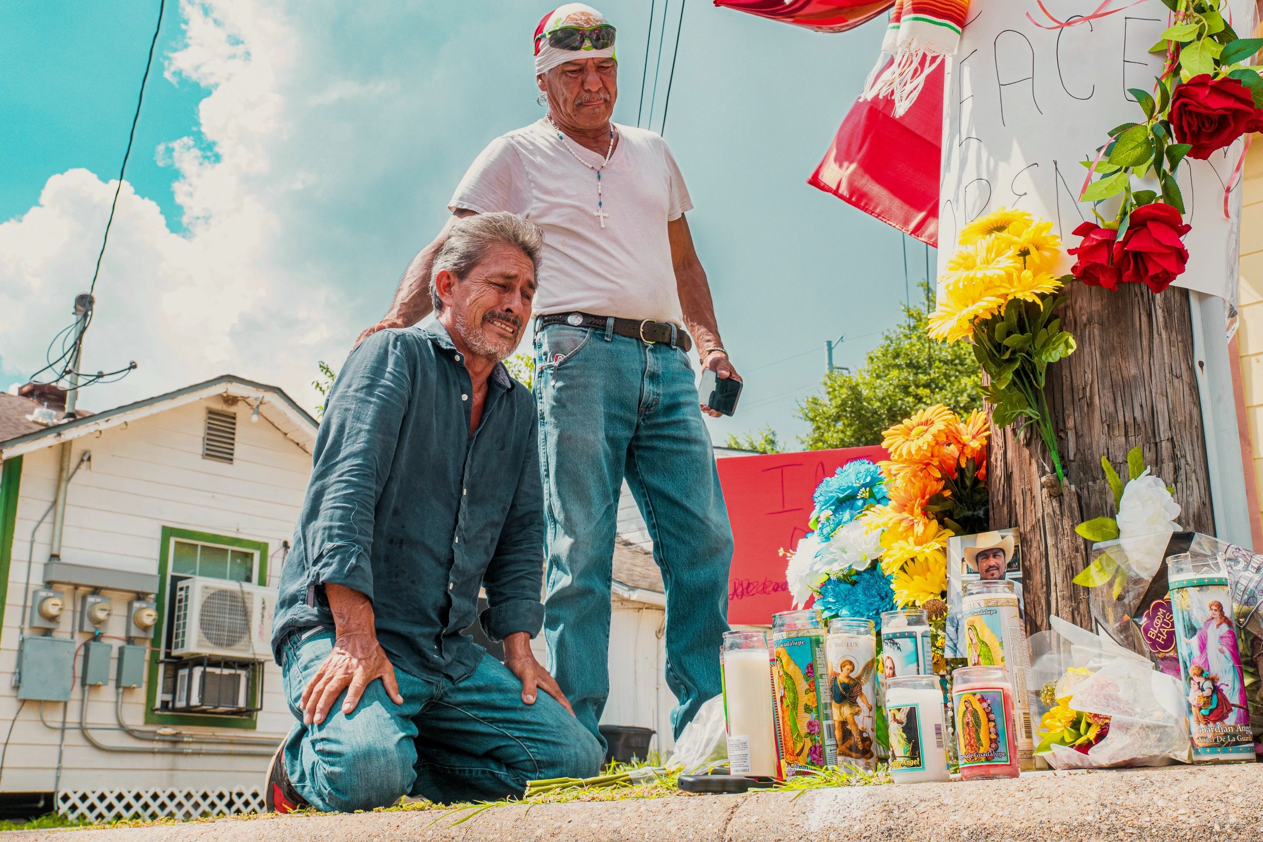 Two men grieving beside a floral memorial.