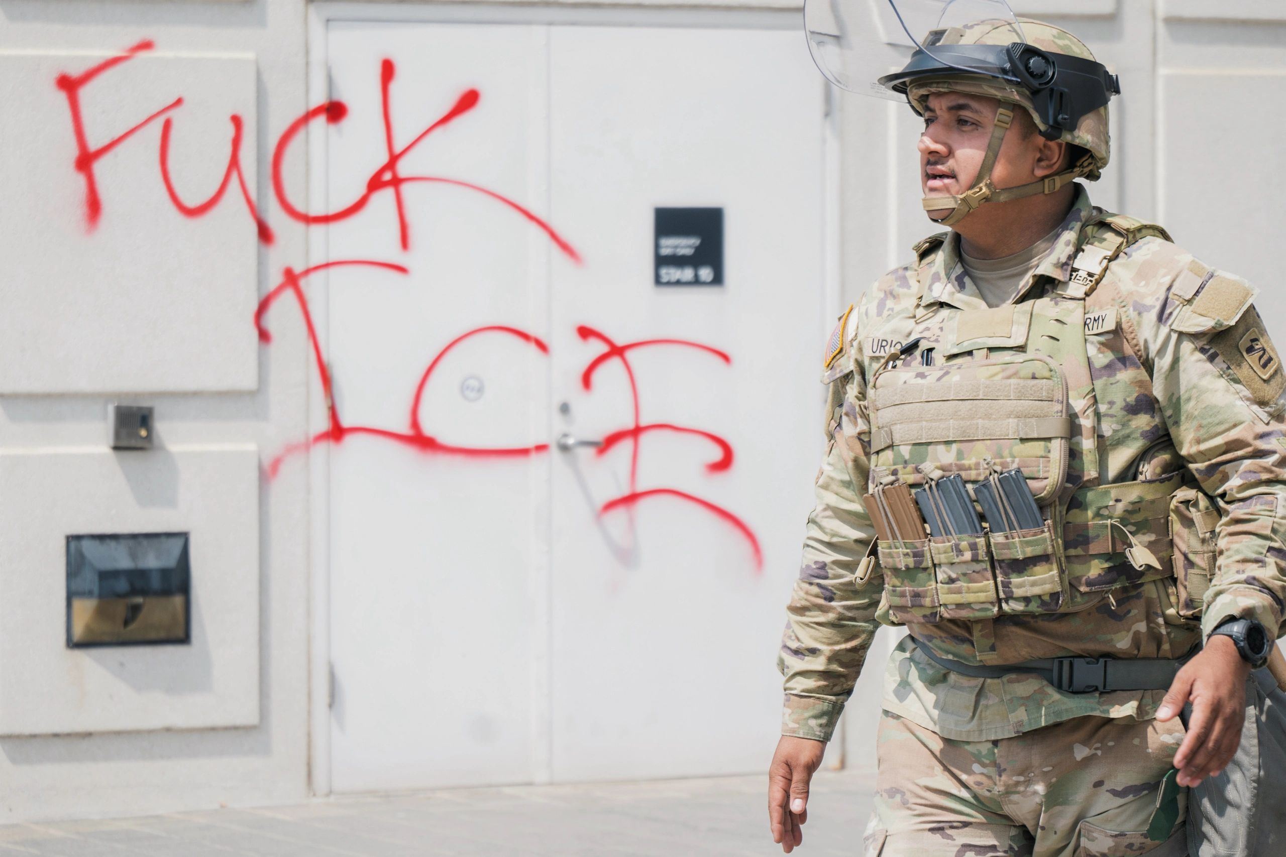 A soldier in camouflage gear walks past a wall with anti-ICE graffiti.