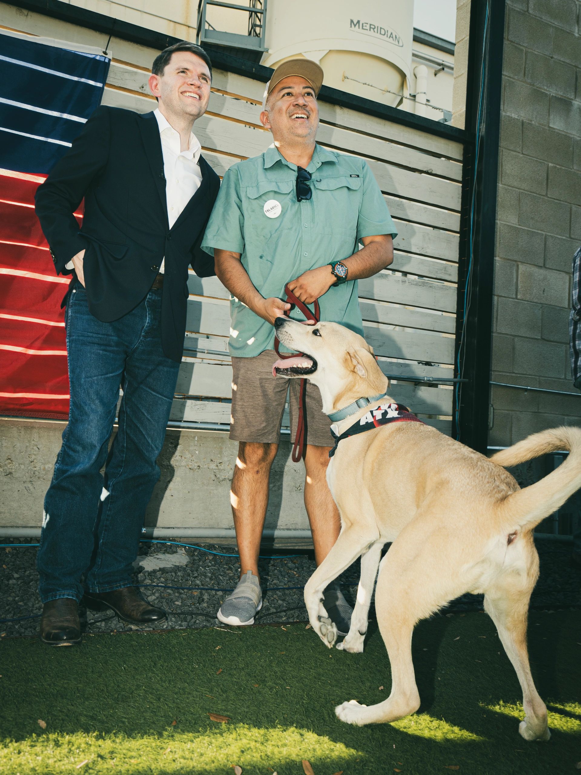 Two smiling men standing outdoors with a happy dog.