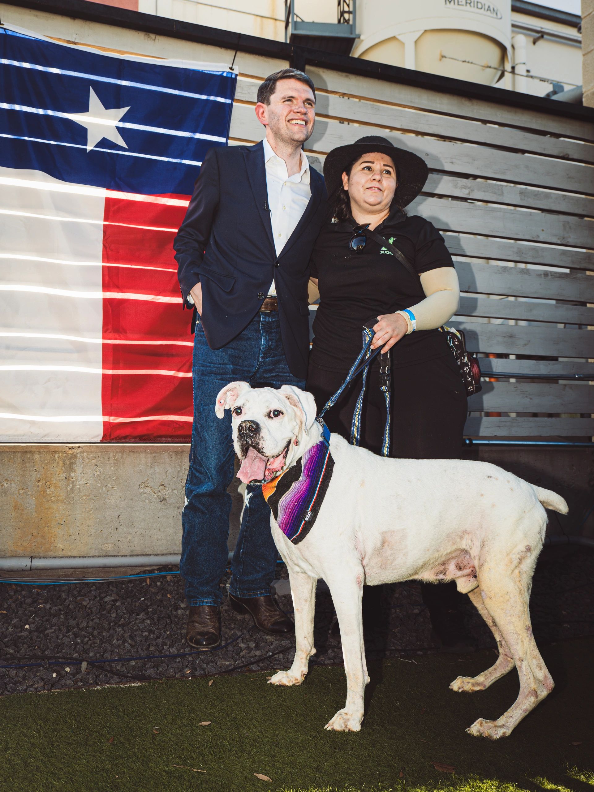 Two people pose with a white dog in front of a Texas flag.