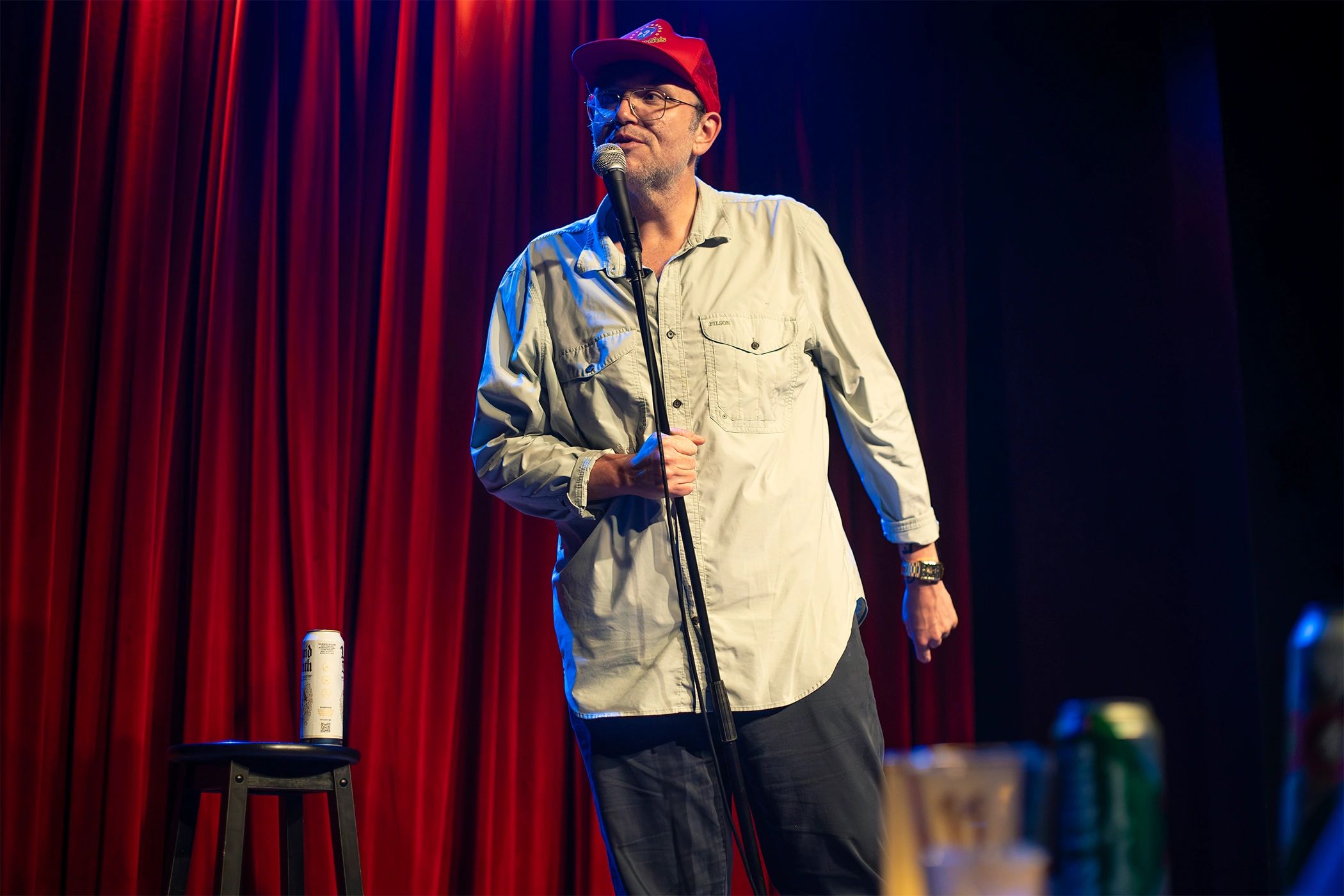 A man in a red cap performs stand-up comedy on stage with a microphone.