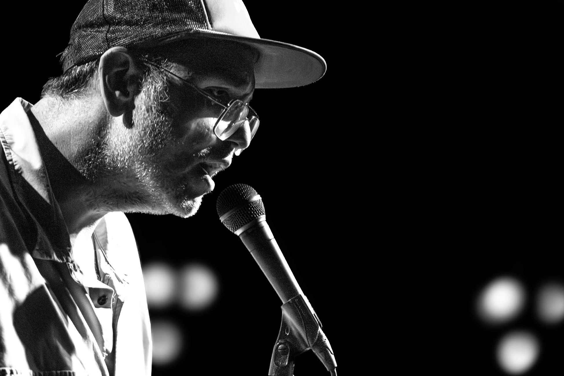 Black and white close-up of a man with glasses and a cap speaking into a microphone.