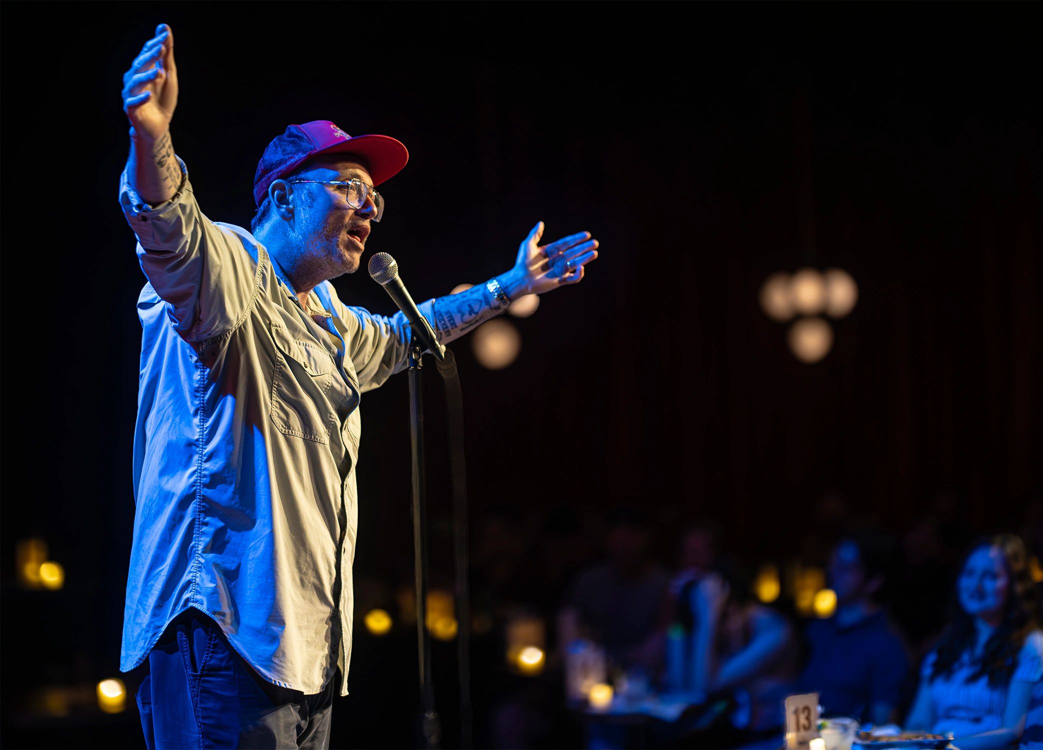 A man passionately performs stand-up comedy on stage with audience in background.