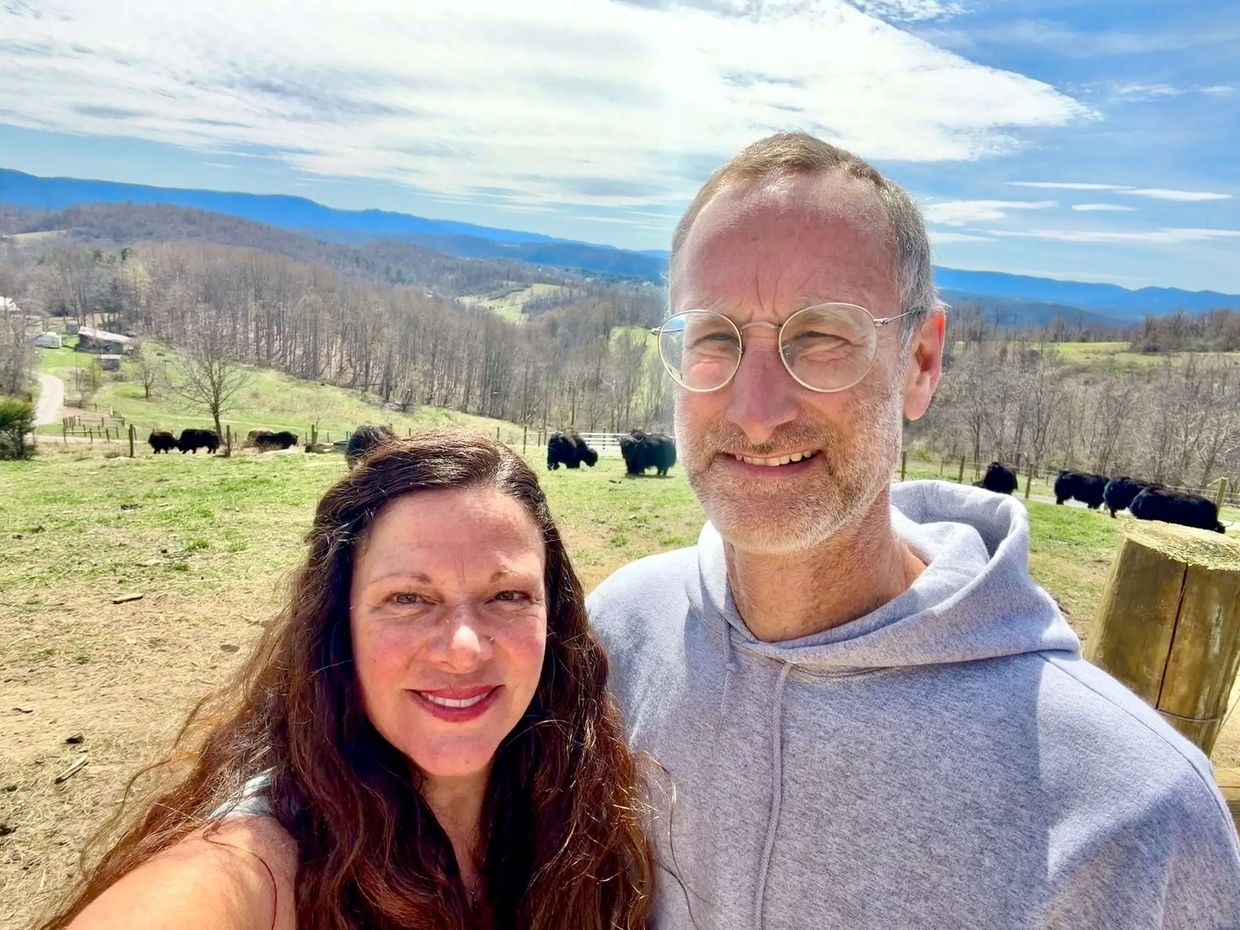 Couple smiling on a farm with black bison and hills in the background.