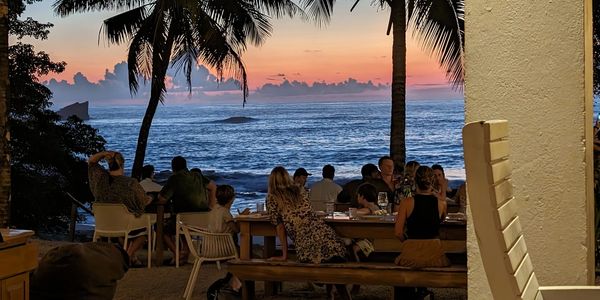 People dining outdoors by the ocean at sunset with palm trees.